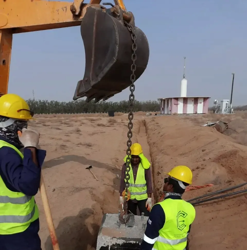 Excavator lowering a concrete block into a trench while crew in Comexperts vests guide it