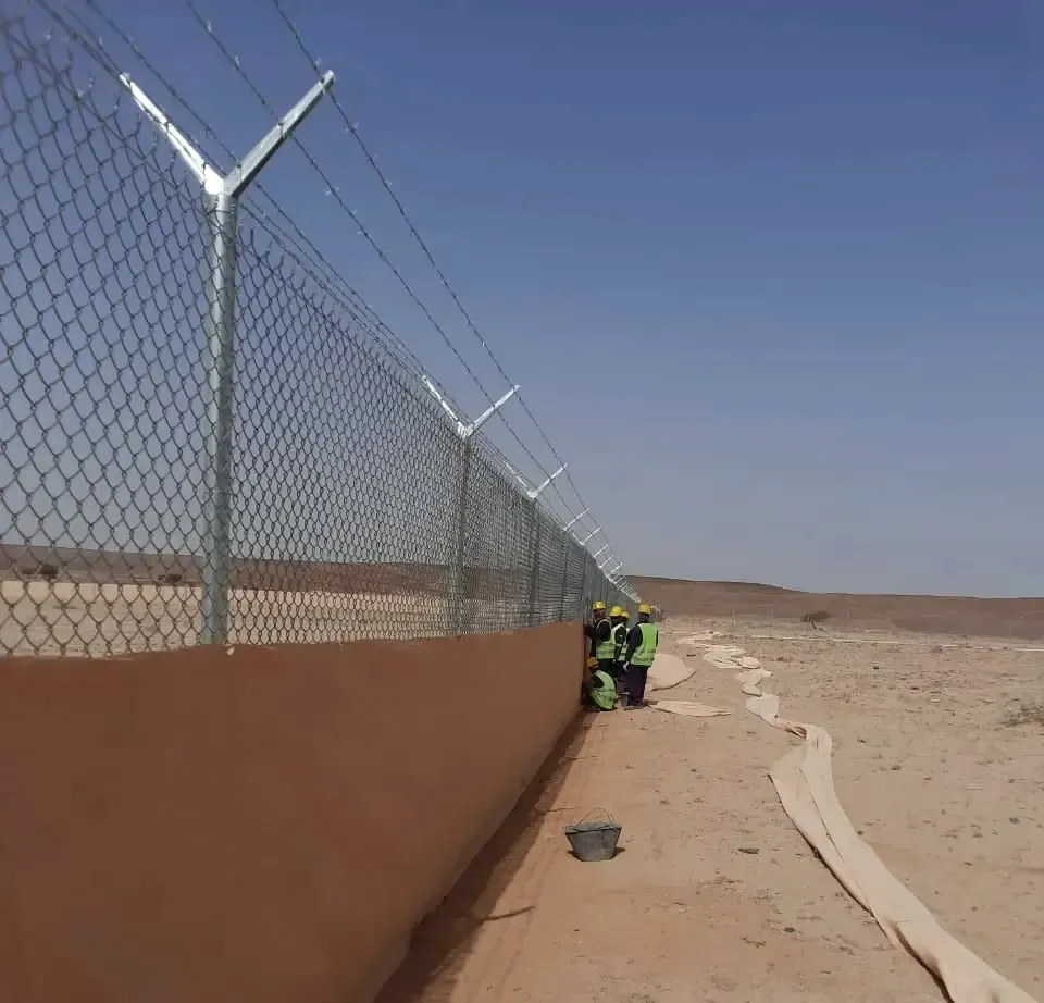 Crew installing a chain-link security fence with barbed-wire arms along a desert perimeter