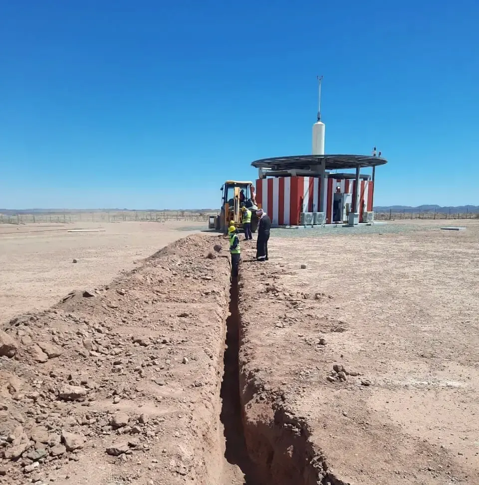 Open cable trench running across a desert site towards an equipment shelter with an antenna